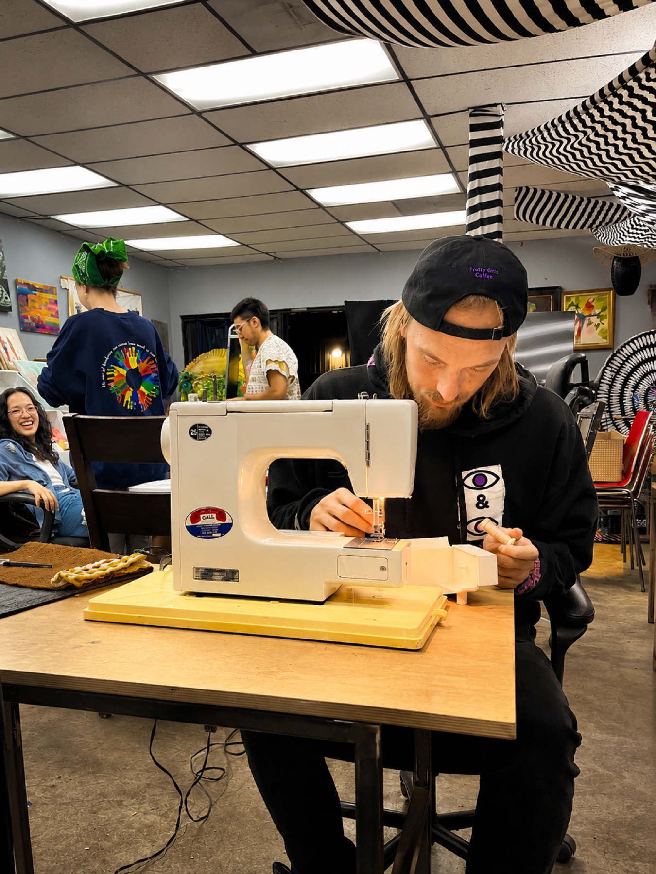 Art Night · at the sewing machine, threading a bobbin while the table laughs behind