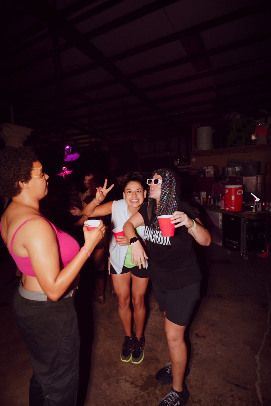Two friends laughing with red cups on the warehouse floor while a third throws a peace sign