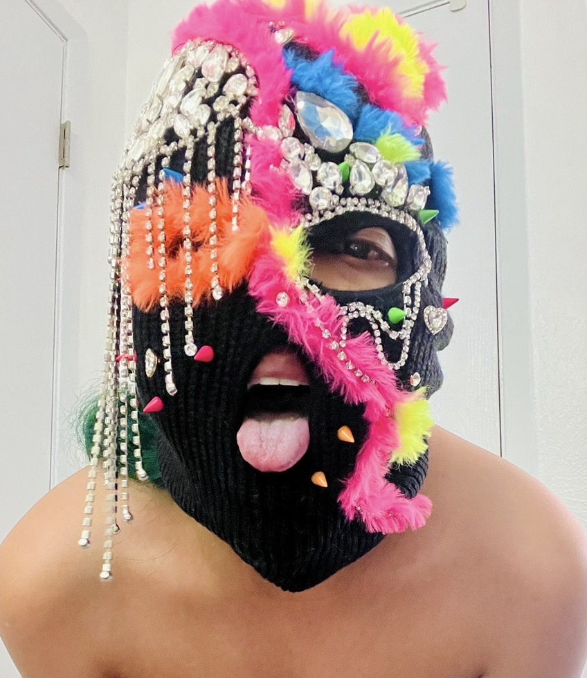 The founder wearing a black Never Normal mask with rhinestone fringe, orange fun fur, and a beaded curtain over one eye