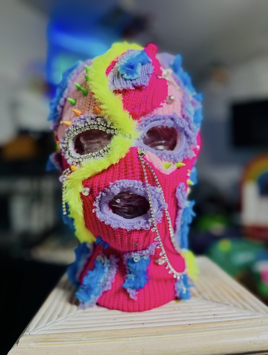 A single Never Normal mask displayed on a stand · pink, lavender, and yellow yarn, rhinestone-ringed eyes, a beaded chain running down the cheek