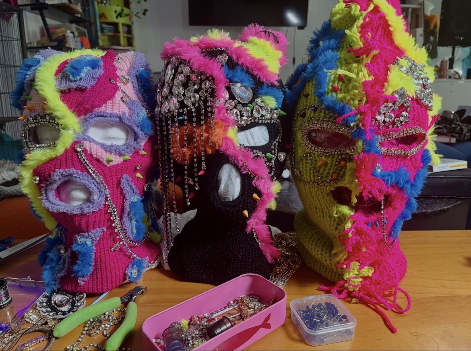 Three Never Normal masks in progress on a workbench, surrounded by pliers, rhinestone chain, and a case of loose gems