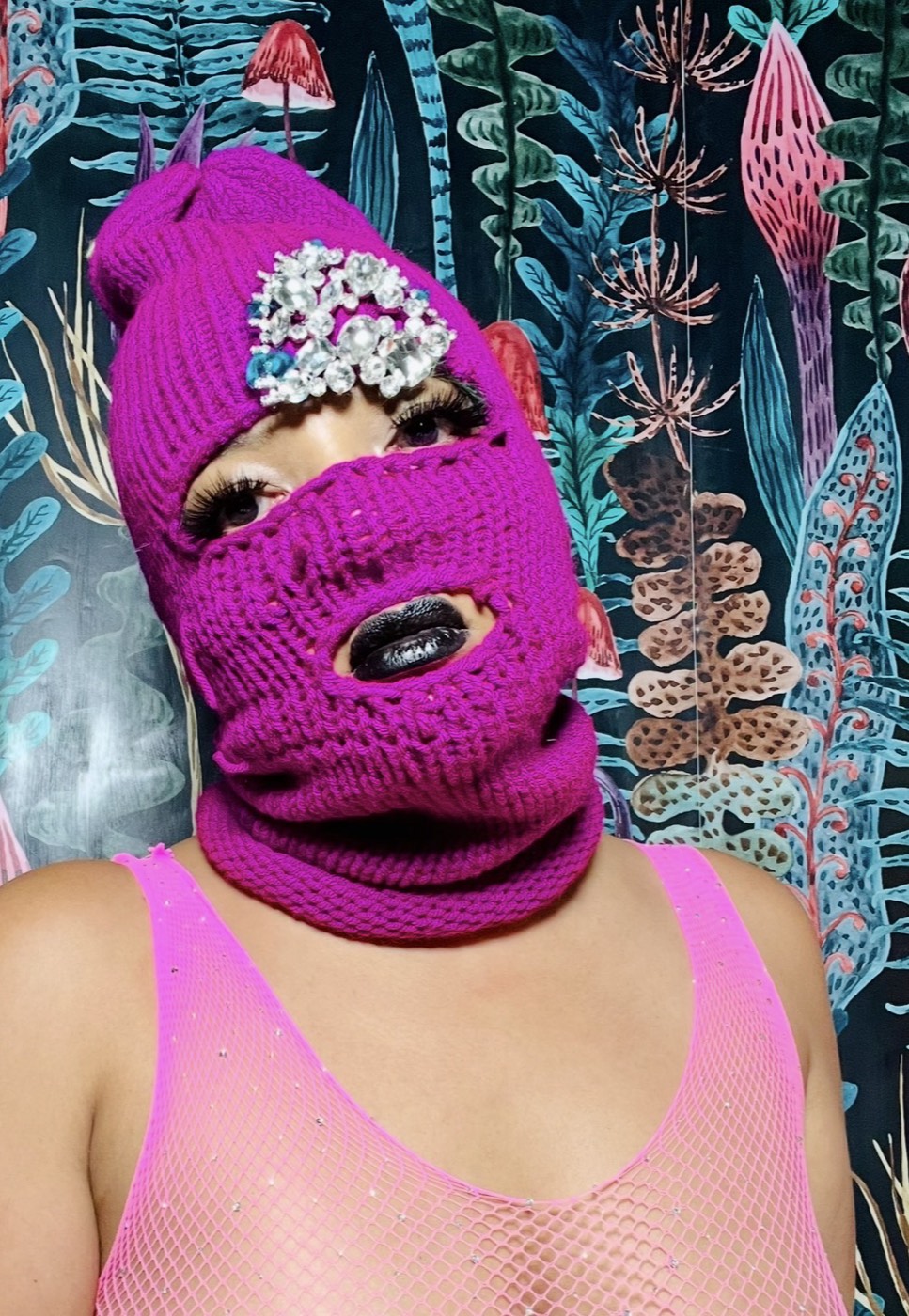 The founder wearing a magenta Never Normal mask with a rhinestone crown detail, against a mushroom-print wallpaper